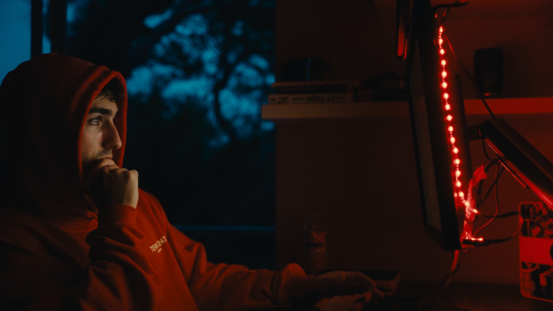 Graded frame: a man in a red hoodie at a desk at night